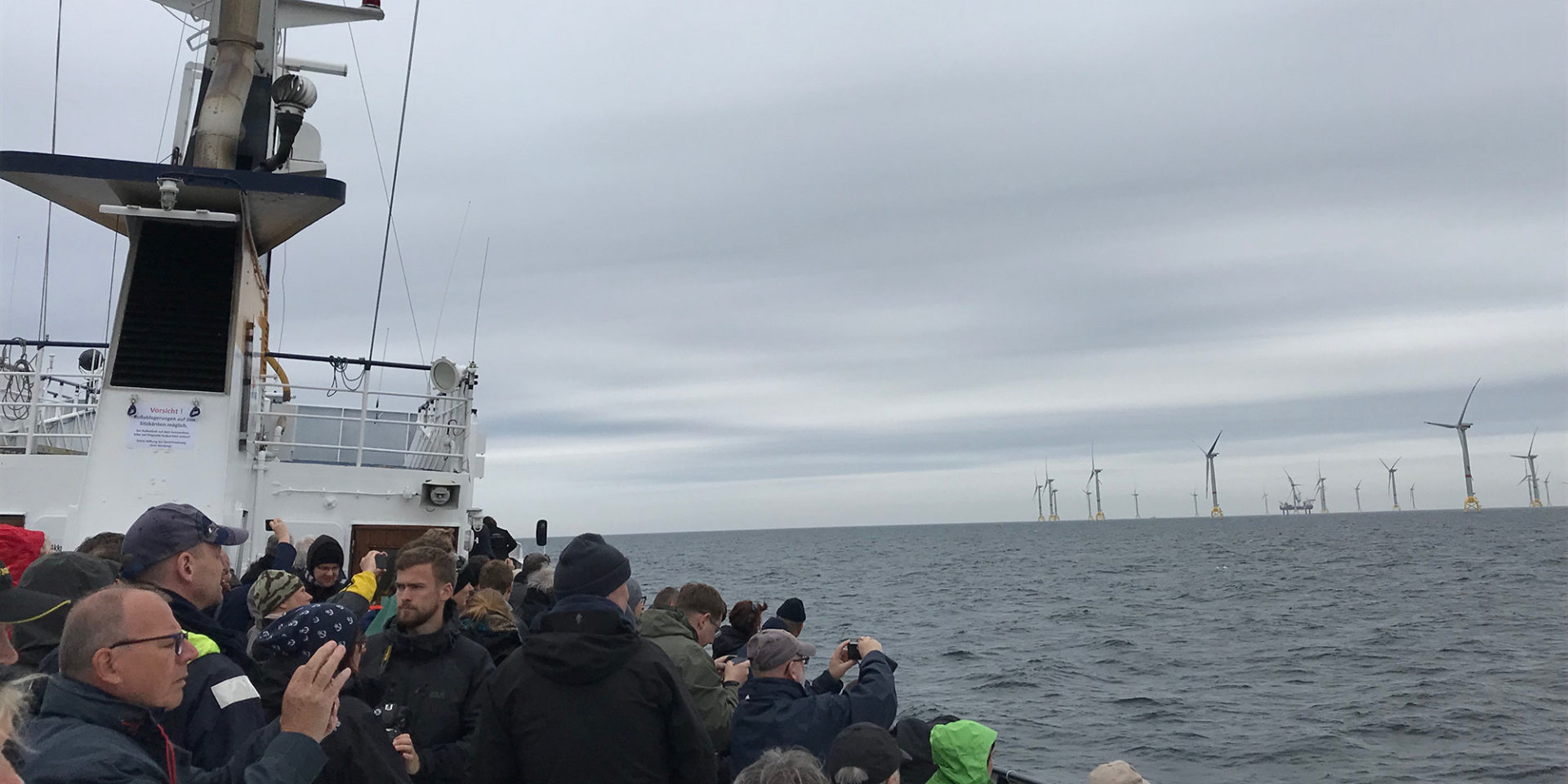 Menschen stehen auf einem Schiff, von dem aus sie einen Blick auf die Windräder in der Ostsee erhalten.