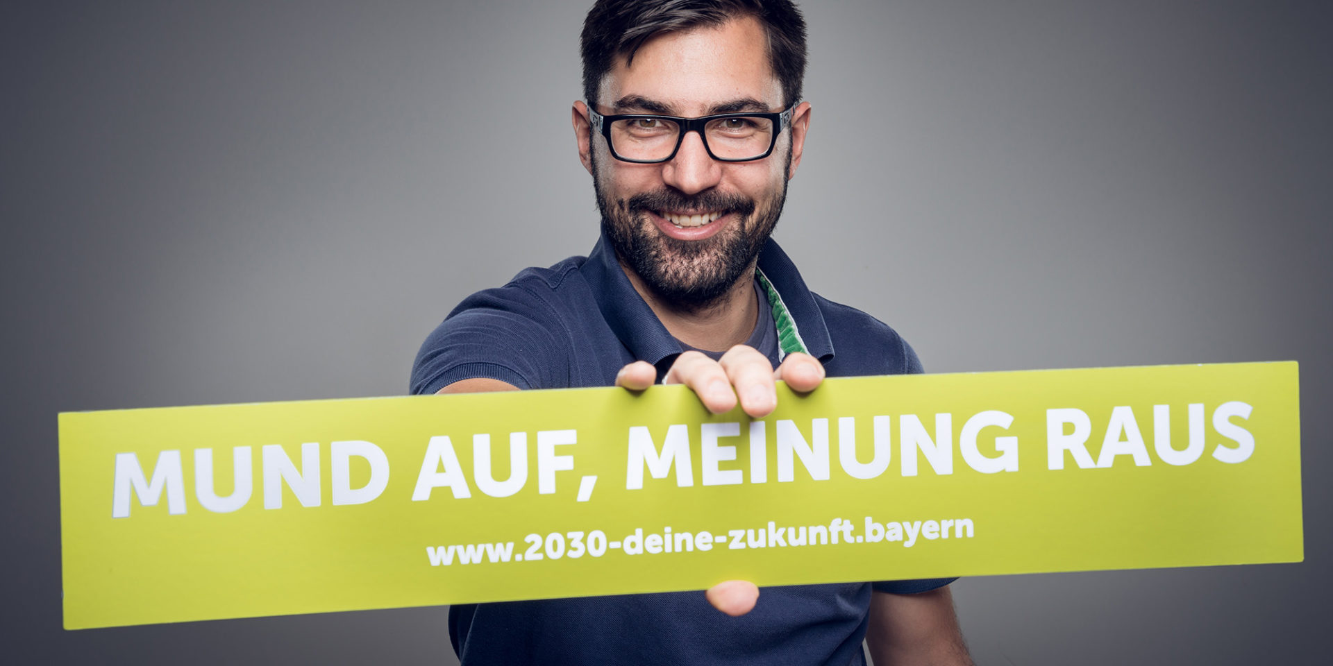 Ein Mann hält einen Banner mit dem Schriftzug "Mund auf, Meinung raus - www.2030-deine-zukunft.bayern" in der Hand