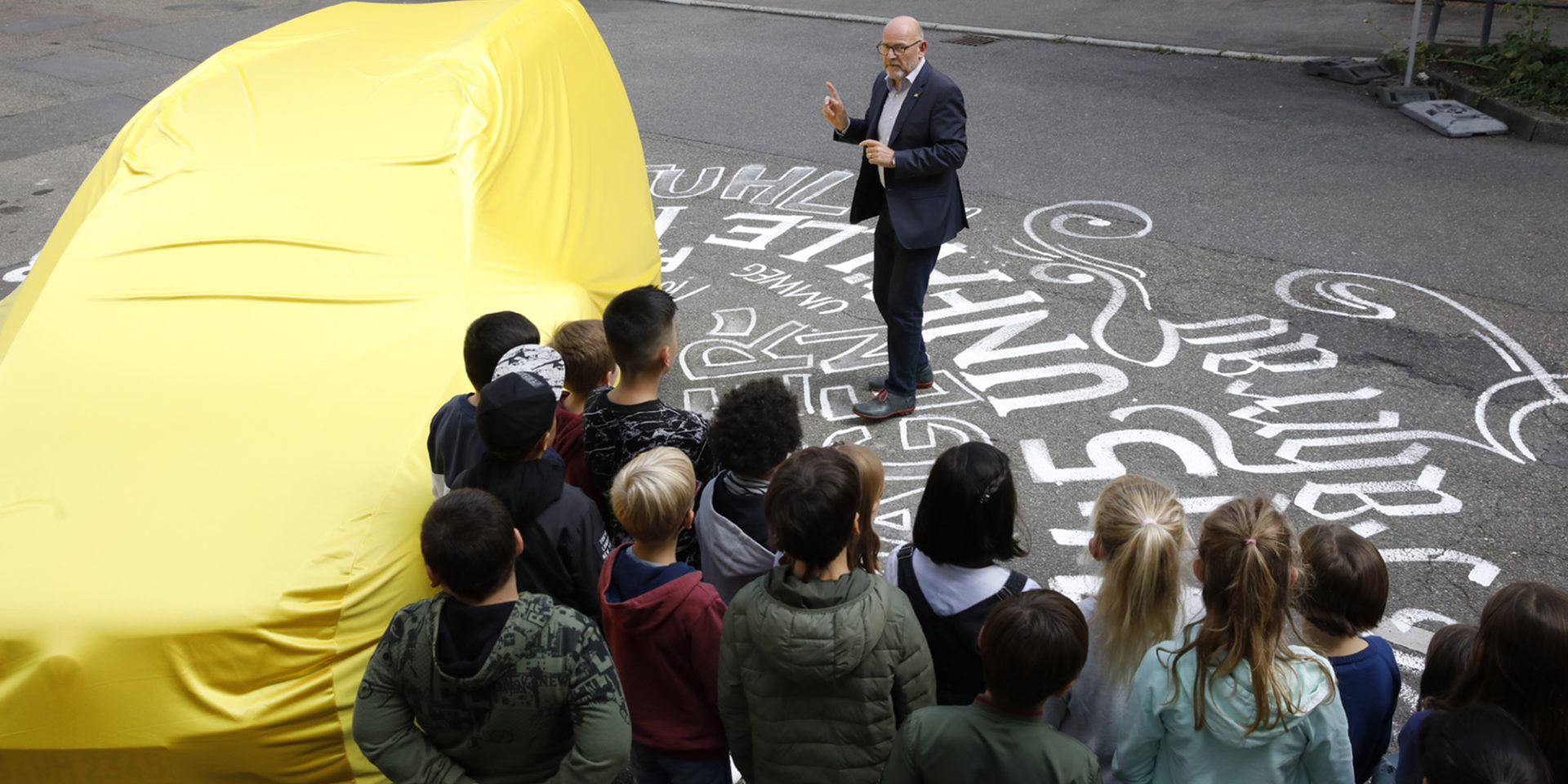 Eine Gruppe von Kindern steht auf einer mit Kreide bemalten Straße und hört einem Mann in Anzug zu - neben der Gruppe ist ein mit einem gelben Tuch verdecktes Auto zu sehen.