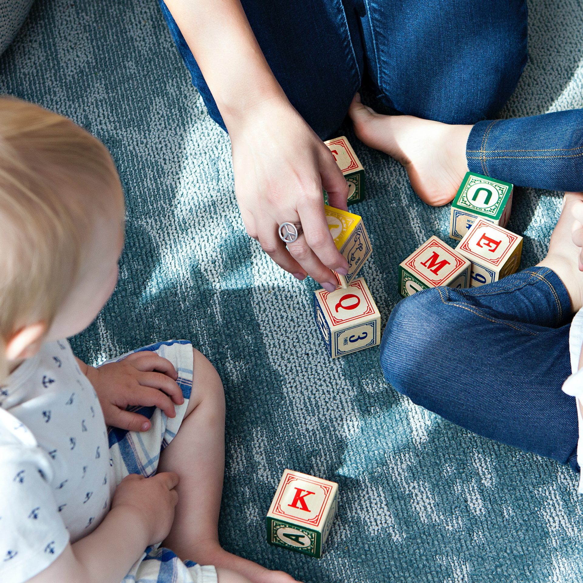 Zwei Kleinkinder spielen auf dem Boden mit Bauklötzen. Die Hände eines Erwachsenen sind zu sehen.