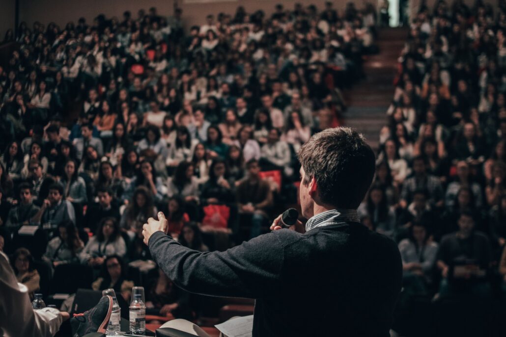 Ein Mann steht mit Mikrofon vor einem Publikum im Hörsaal und spricht mit anderen Personen auf der Bühne im Rahmen einer Podiumsdiskussion.