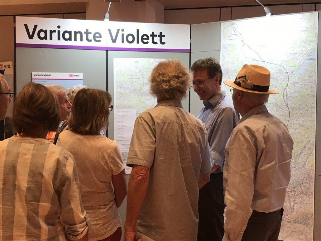 Eine Gruppe von mehreren Personen spricht mit einem Mann im Profil. Sie stehen direkt vor einer Stellwand mit der Überschrift „Variante Violett“, zwei Karten und einer Infotafel.