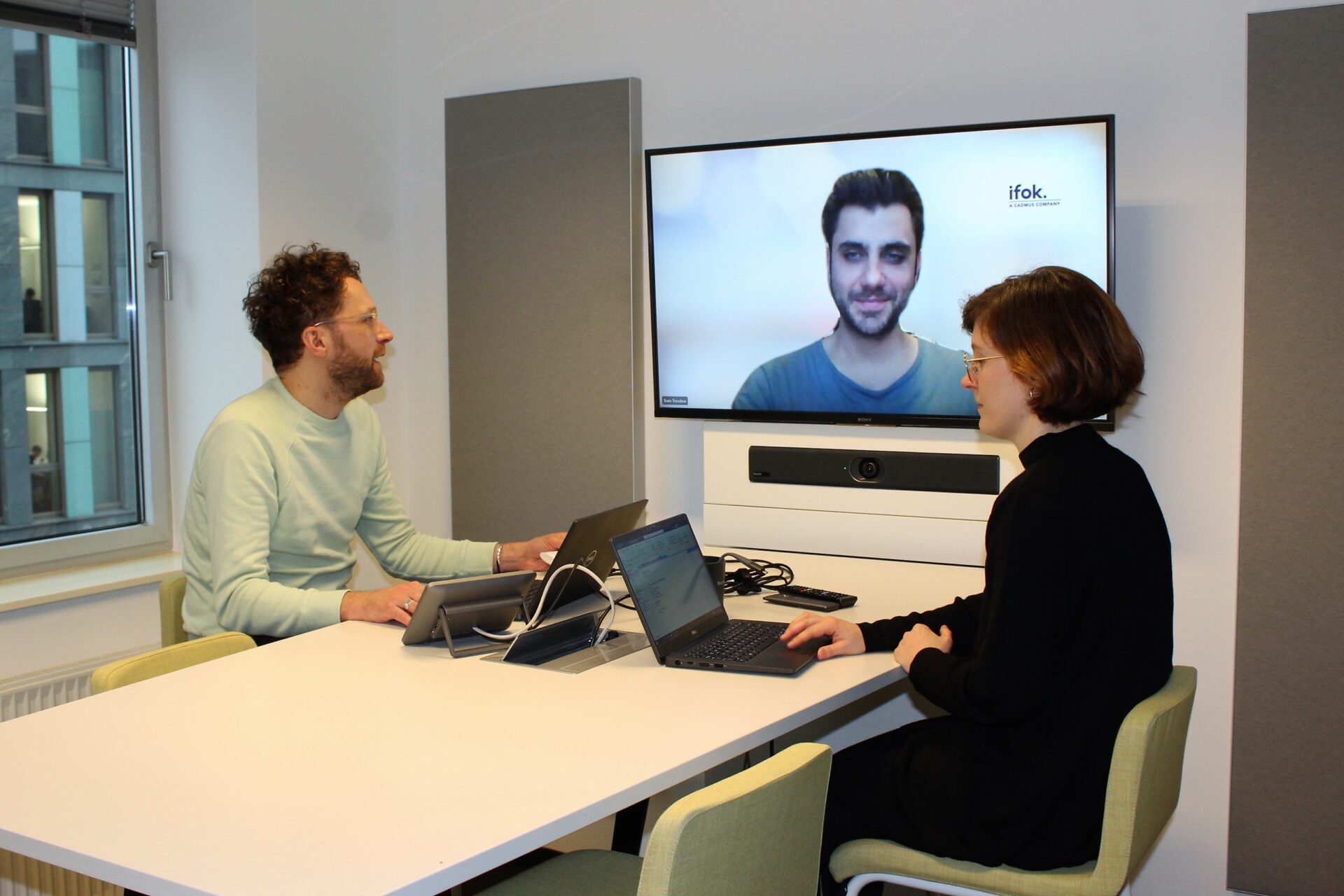 Zwei ifox arbeiten in einem Besprechungsraum, ein Kollege ist digital zugeschaltet.