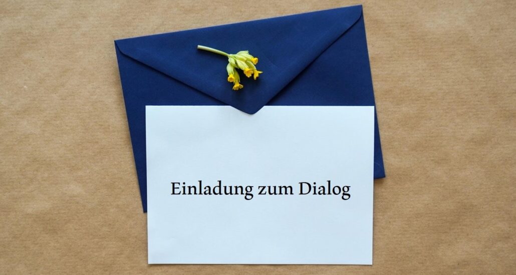 Dunkelblauer Briefumschlag und weiße Karte mit Schriftzug "Einladung zum Dialog" liegen auf braunem Packpapier.